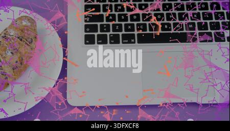 Showing silver laptop, black keys and pad on purple mat, croissant on plate, pink-orange confetti Stock Photo