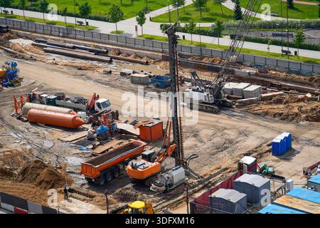Workers operate heavy machinery at a busy construction site, utilizing cranes and trucks to move materials surrounded by green spaces under a clear bl Stock Photo