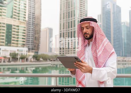 Middle Eastern businessman in traditional dress stands in modern city, engaged in planning and investment using a tablet for future success. Stock Photo