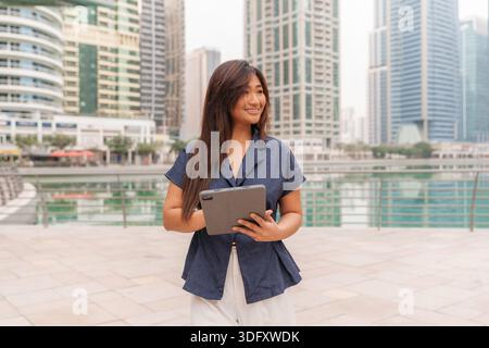 Asian woman stands outdoors in a smart city, engaging with her tablet while doing real estate business. Stock Photo