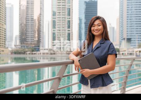 Asian woman stands by a railing in a smart city, using a tablet while embracing urban life and digital work culture. Stock Photo