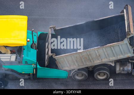 Asphalt paver lays down an even layer of hot new asphalt from the back of a truck during road repairs, top view. Stock Photo