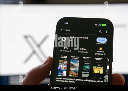 Man holding phone with xAI app chatbot Grok install screen on Google Play, with the X logo behind. Concept: AI generated images, manipulated images Stock Photo