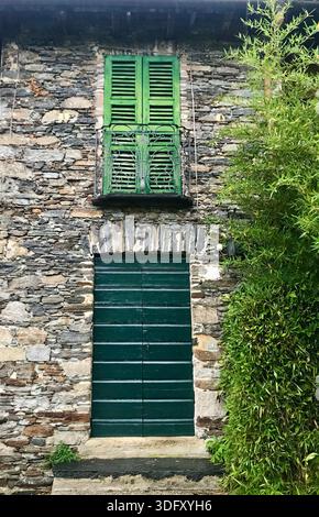 A unique house facade in Orta San Giulio, Italy, featuring a textured stone wall, green closed shutters, and a towering tree beside the building. Stock Photo