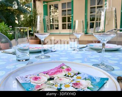 Spring-inspired table setting arranged outdoors in the garden of a French country house with flowered napkins and elegant tableware. Stock Photo