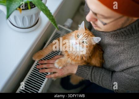 Man cuddling a fluffy ginger cat by a radiator, finding cozy warmth and comfort at home while coping with high energy costs and the rising cost of living Stock Photo