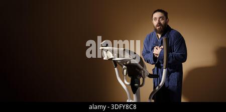 Young man in bathrobe looking surprised while using a stationary exercise bike against a brown studio background. Stock Photo