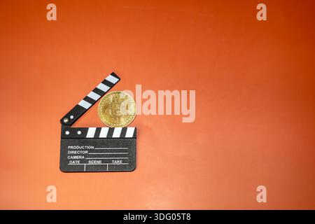 A Bitcoin coin rests on a film clapperboard against an orange background. This image suggests cryptocurrencies' potential in film production. Stock Photo