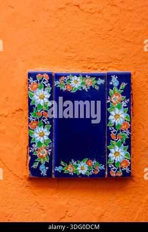 A stunning blue ceramic tile adorned with floral designs contrasts beautifully with the vivid orange wall, creating a striking visual display. Stock Photo