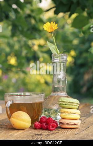 Ripe fruits of apricot and strawberries, calendula flower with a stem in a glass flask, macaroons and cup of green tea on wooden boards with blurred n Stock Photo