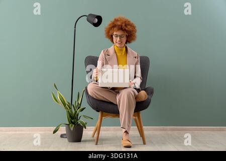 Redhead female freelancer working with laptop in armchair near green wall Stock Photo