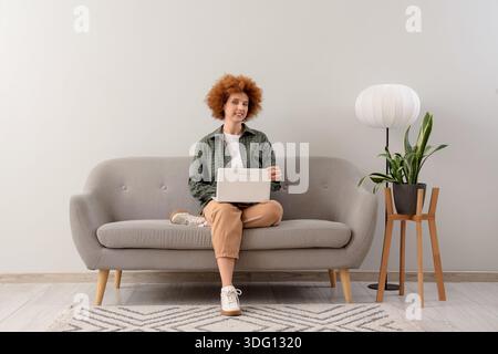 Redhead female freelancer working with laptop on sofa near light wall Stock Photo
