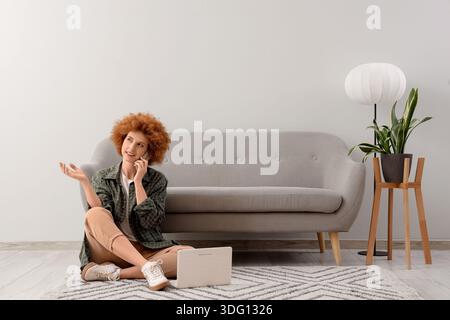 Redhead female freelancer with laptop talking by mobile phone near light wall Stock Photo