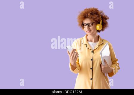 Redhead female freelancer in pajamas with gadgets on lilac background Stock Photo