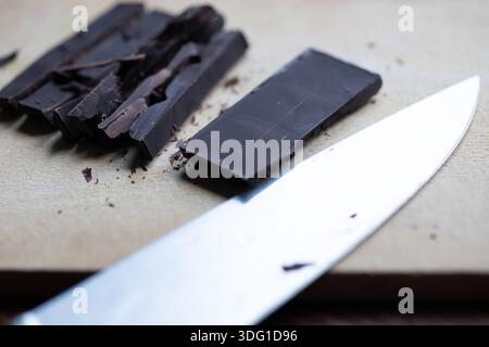 Close-up of a sharp knife chopping a rich dark chocolate bar on a wooden board Stock Photo
