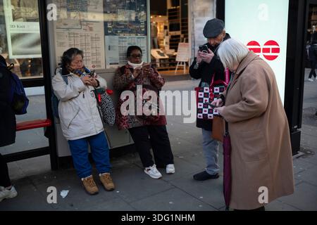 Silver surfers, elderly and middle aged commuters checks their smartphones whilst waiting at a bus stop in London's West End, England, United Kingdom. Stock Photo