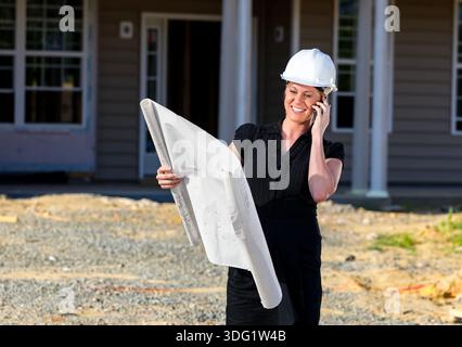 On a sunlit build site, a female engineer reviews blueprints, checking measurements and details as framing rises around her, guiding the construction Stock Photo