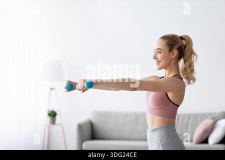 Happy caucasian pretty young blonde lady lifting dumbbells at home in domestic gym, side view Stock Photo