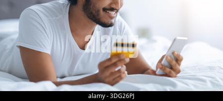 A Man is Lying on a Bed and Smiling as He Uses His Smartphone Stock Photo