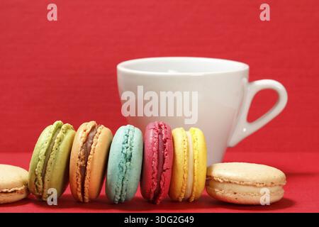 Sweet macarons cakes of different color with white porcelain cup on red textured surface and background Stock Photo