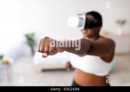 Virtual training. Overweight black lady in VR glasses using fighting simulator, making punch at home, focus on fist Stock Photo