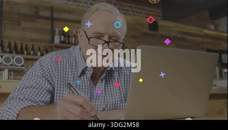 Writing senior man wearing checked shirt, reading glasses, using laptop and noting with pen at cafe Stock Photo