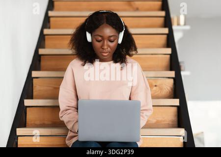 Black Female Working On Laptop Wearing Headphones Sitting At Home Stock Photo