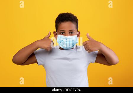 Funny black boy wearing protective face mask, showing thumb ups Stock Photo