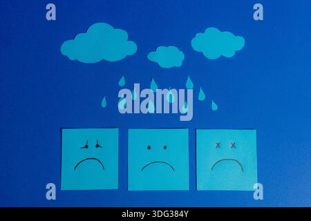 Composition on a blue background featuring three sad face sticky notes under paper cutout clouds and raindrops. Stock Photo