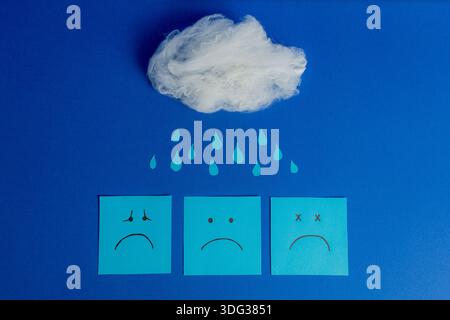 Blue Monday visual concept featuring a cotton cloud, paper raindrops, and three blue notes with various sad expressions. Stock Photo