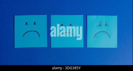 Three square sticky notes arranged in a row on a blue background, showing different expressions of sadness, tiredness, and depression. Stock Photo