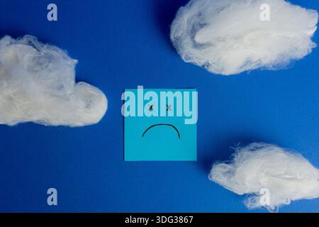 Minimalist composition on a blue background featuring a sad face sticky note surrounded by three white cotton clouds. Stock Photo