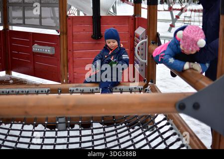 Two toddlers and dad playing together on a modern playground bridge in winter. Children wearing warm colorful snowsuits explore balance and coordinati Stock Photo