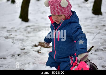 Toddler girl kneeling in the snow and playing with a snowball in a winter park. Child wearing warm colorful snowsuit explores nature, textures, and se Stock Photo