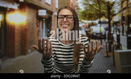 Woman with glasses beckons with both hands on a busy city street, smiling broadly; welcoming playfulness casual. Stock Photo