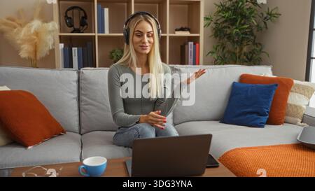 Young woman wearing headphones gestures to laptop in living room at home; virtual meeting comfort learning. Stock Photo