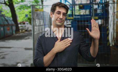Man wearing glasses and dark shirt with wedding ring raises right hand palm out and places left hand on chest in street near metal cages and dumpsters Stock Photo