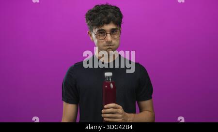Man young hispanic holding a bottle with hand and cheeks puffed in purple studio wearing glasses and black shirt; playful curiosity. Stock Photo