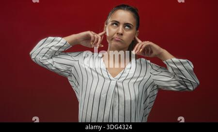 Woman plugging ears with fingers in studio against a deep red backdrop, wearing a white striped blouse and looking upward with hands raised; frustrati Stock Photo