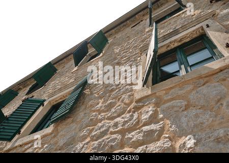 Low angle view of traditional stone house facade with green shutters and blank white sky, textured wall background with copy space. Stock Photo