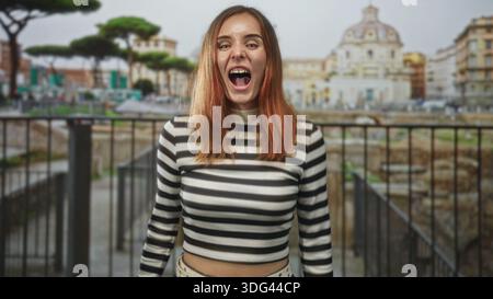 Young redhead woman with mouth open screaming, eyes closed, leaning on a railing at a historic building; playful surprise. Stock Photo