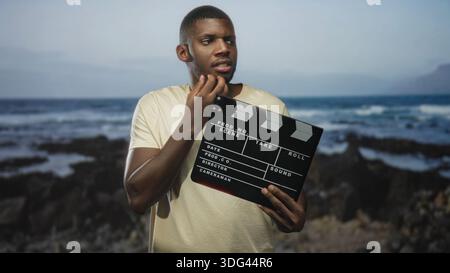 Man holding clapperboard, stroking chin and looking aside on rocky beach shore with waves and volcanic rocks; thoughtful filmmaking inspiration. Stock Photo