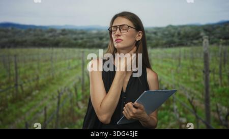 Woman holding clipboard with hand on throat and throat pain while inspecting vineyard rows in forest; discomfort crop inspection. Stock Photo