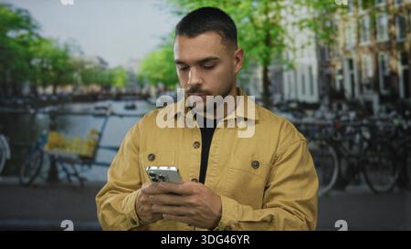 Man in mustard jacket holds smartphone and scrolls screen on street beside canal with parked bicycles and brick buildings; quiet reflection. Stock Photo