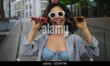 Woman wearing bikini holds multiple sunglasses while smiling on a street with modern buildings, embodying fashion and joy in an urban outdoor environm Stock Photo