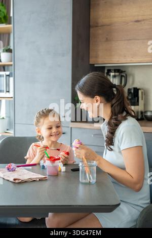 Mother and daughter sitting at table at home and decorating Easter eggs together. Family holiday tradition, creative bonding activity and warm Easter Stock Photo