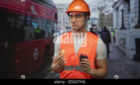 Man engineer in orange hardhat and safety vest holding coffee cup and pastry on a busy street; work break content. Stock Photo