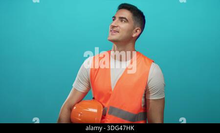 Man holding orange hard hat and wearing orange safety vest in studio against turquoise backdrop; safety professionalism optimism. Stock Photo