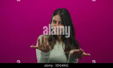 Hispanic woman beckons with open hands in a pink studio with a bright friendly smile; warm invitation. Stock Photo