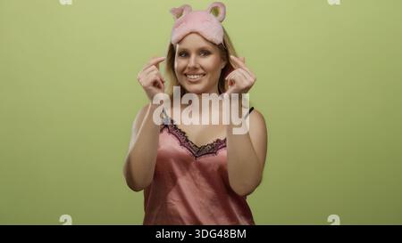 Young blonde woman in pink nightwear makes heart gesture with hands against yellow background wearing a playful sleeping mask, conveying a sense of af Stock Photo